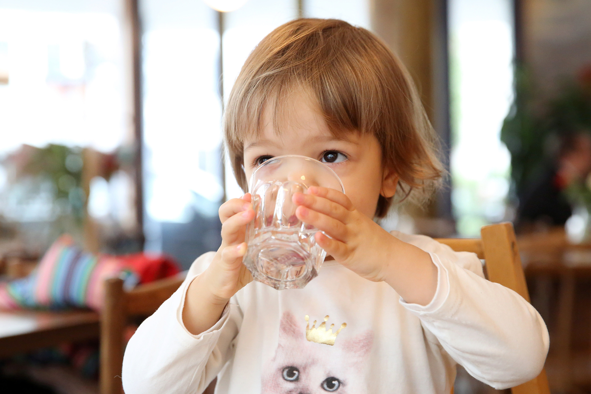 Ein Kleinkind trinkt Wasser. Es führt ein Glas mit zwei Händen an den Mund