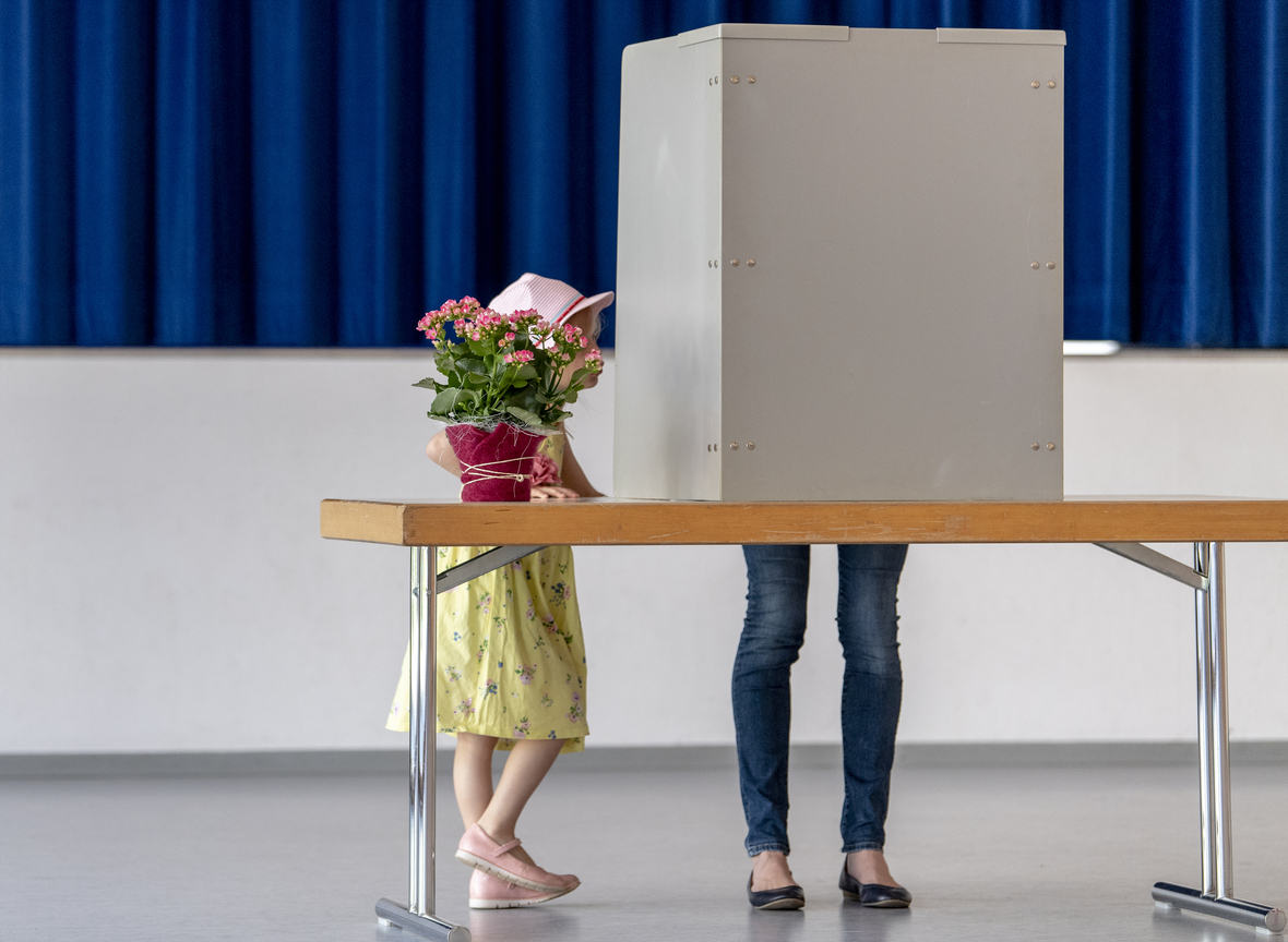 Ein kleines Mädchen steht neben einer Wahlkabine, in der die Mutter den Stimmzettel ausfüllt.