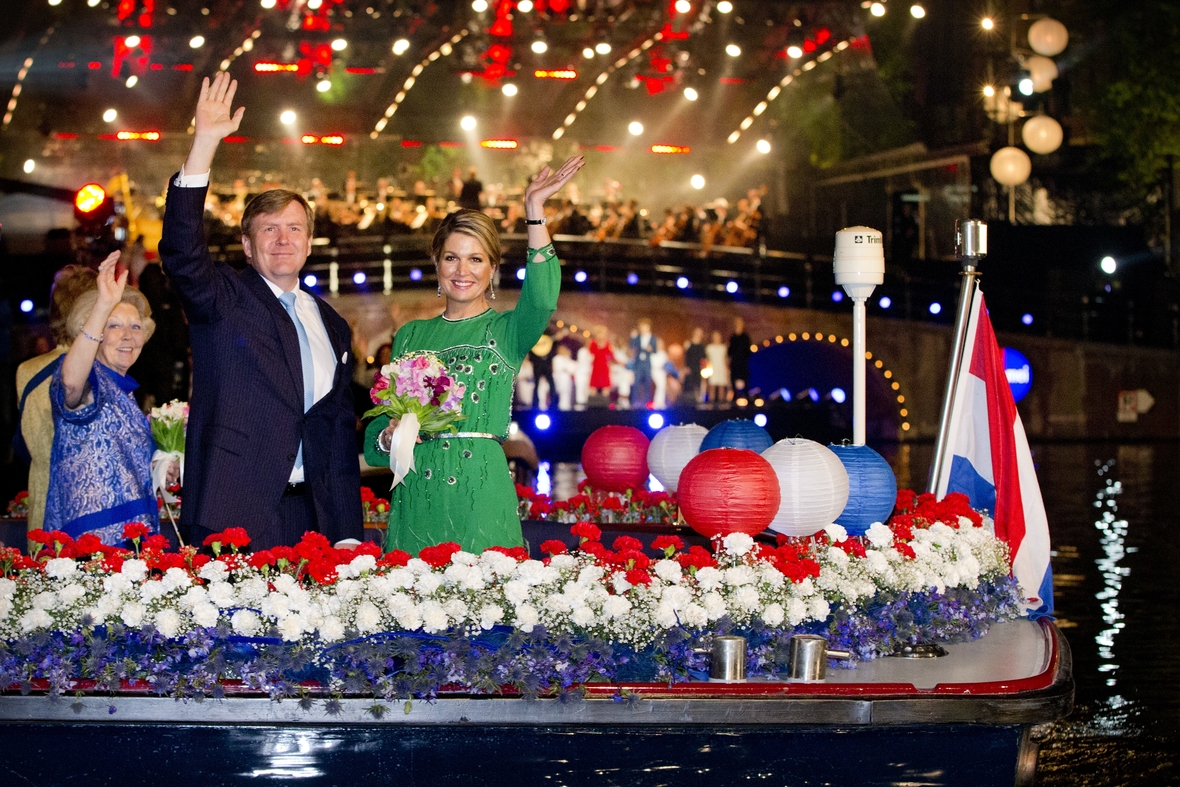 Das Foto zeigt den niederländischen König Willem-Alexander, seine Frau Königin Maxima und Prinzessin Beatrix der Niederlande. Sie stehen winkend auf einem Boot, das in den Nationalfarben der Niederlande, also in Rot, Weiß und Blau, dekoriert ist.  