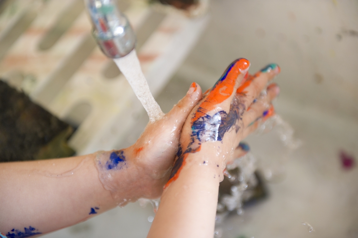Ein Junge wäscht sich seine mit Farbe verschmierten Hände unter einem Wasserhahn, mit denen er zuvor ein Bild gemalt hat.