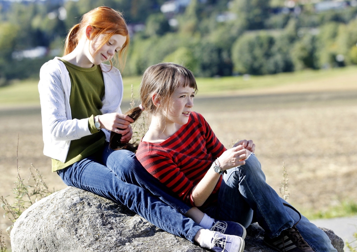 Szenenbild von links nach rechts: Beate auf einem Stein sitzend spielt mit dem Pferdeschwanz ihrer Freundin Anne