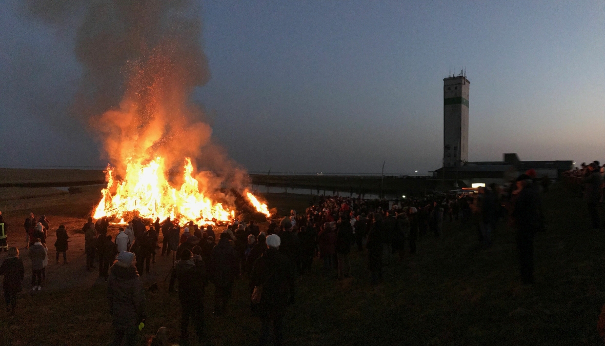 Menschen stehen um ein hoch loderndes Feuer. Gezeigt wird das traditionelle Biike-Feuer, das an den Küsten Nordfrieslands den Winter verabschiedet.