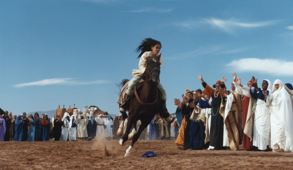 Szenenbild: Das Mädchen Zaina, in der Mitte des Bildes, galoppiert auf ihrem Pferd Zingal an den jubelnden Zuschauern vorbei.