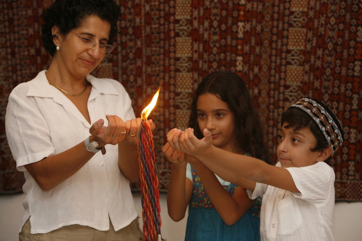 Eine Mutter mit ihren zwei Kindern beim Schabbat. Die Mutter und Kinder stehen um eine entzündete Kerze.