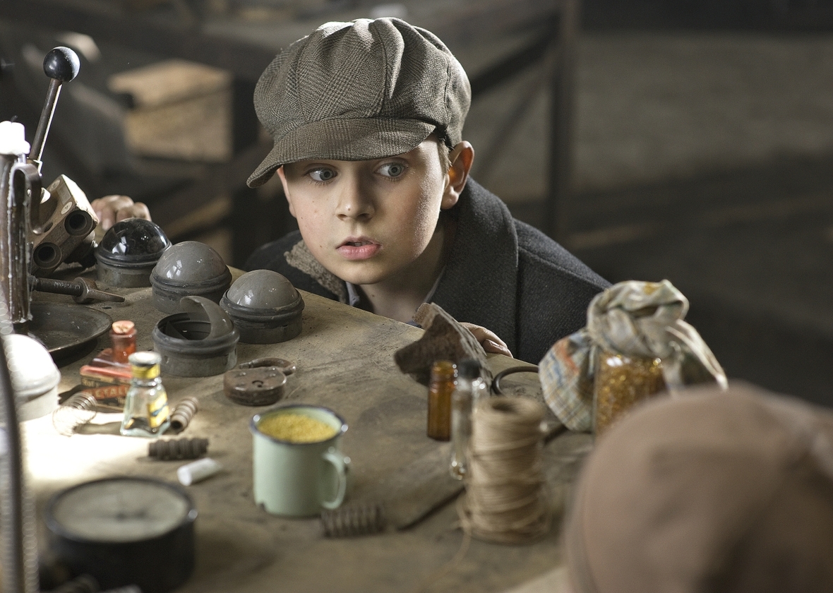 Szenenbild: Der Junge Alexander mit einer Kappe auf dem Kopf entdeckt auf einem Tisch voller Gegenstände die Perlmutterfarbe